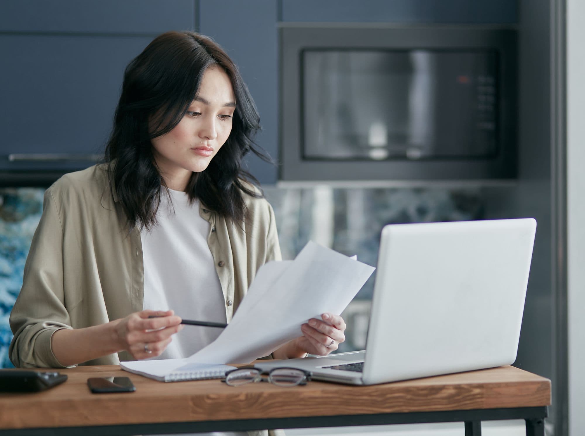 A woman at her kitchen table reading through printed papers beside her laptop