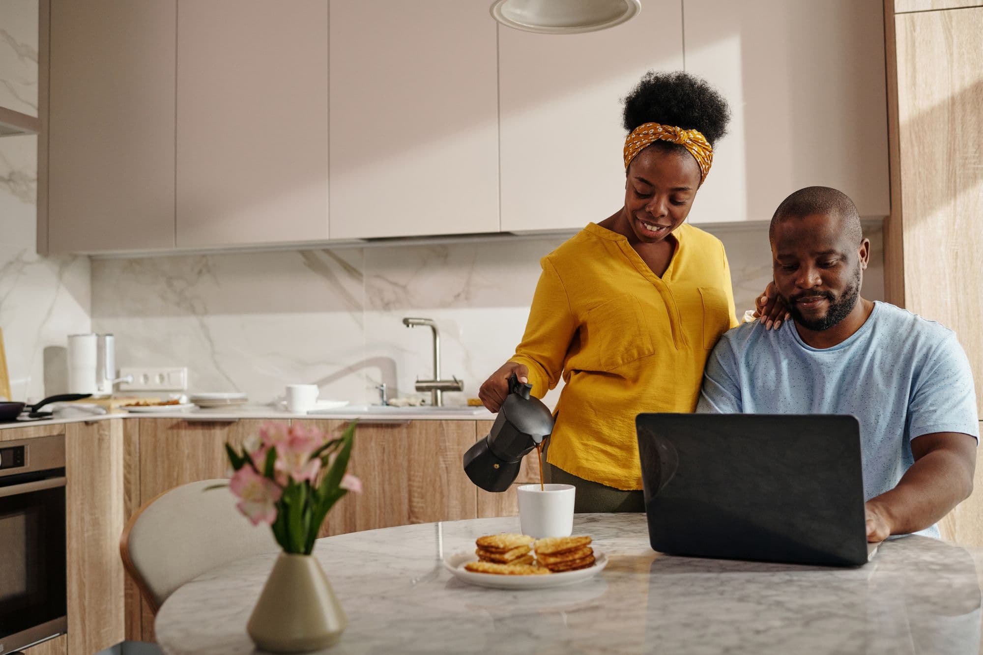 A couple at their kitchen table at breakfast, one pouring coffee while the other looks at a laptop