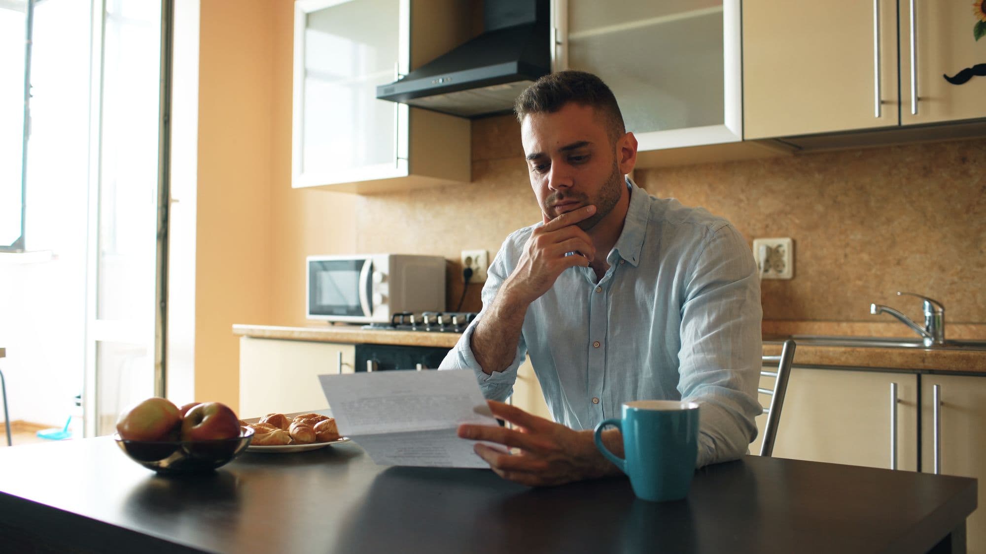 A man at his kitchen table reading a letter closely, with a mug of tea beside him